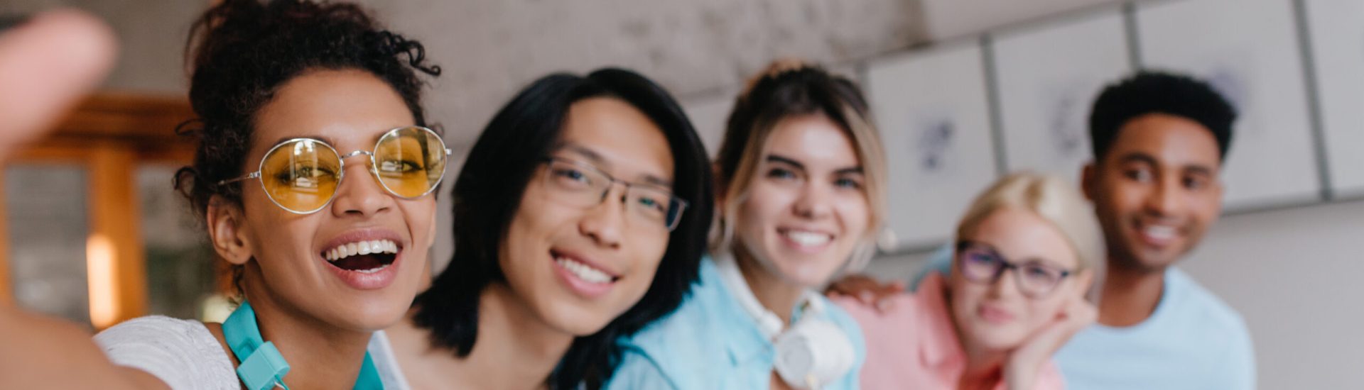 Inspired girl in stylish yellow glasses making selfie with her asian university friend and other students. Charming young woman with light-brown skin taking photo of herself with people on background.