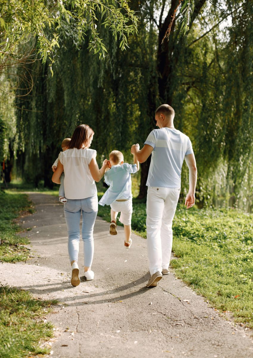 Mother, father, older son and little baby daughter walking in the park. Family wearing white and light blue clothes. Mother holding daughter on a hands.